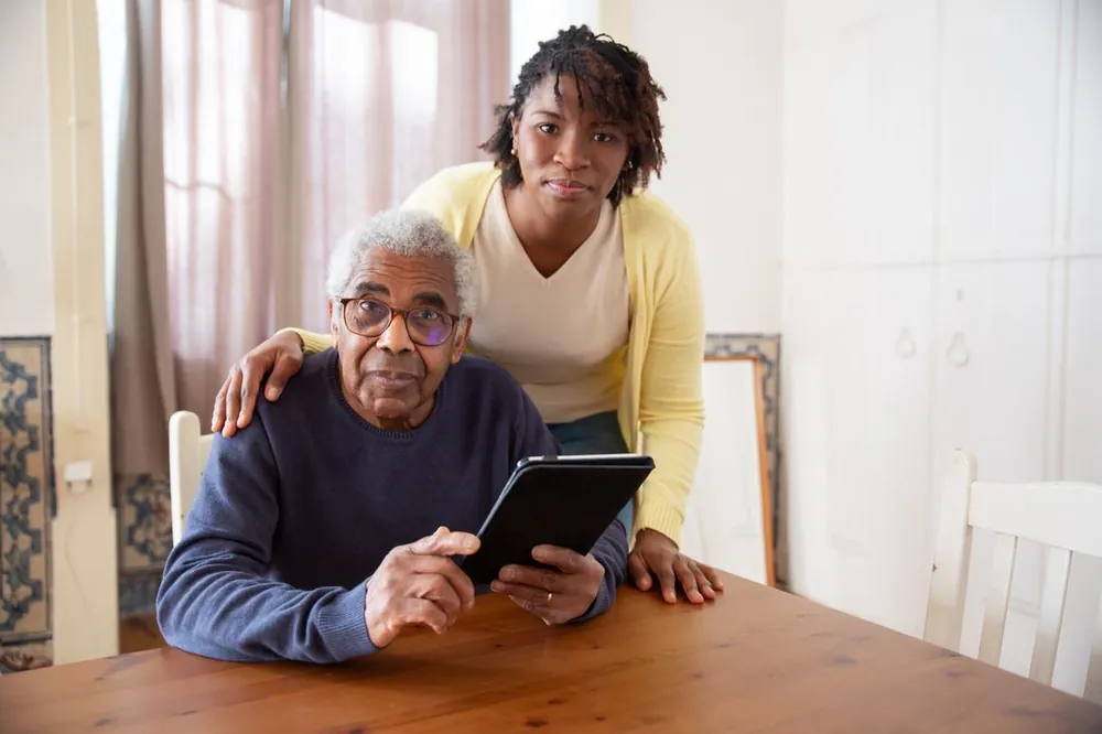 A carer standing supportively beside an older man seated at his table at home