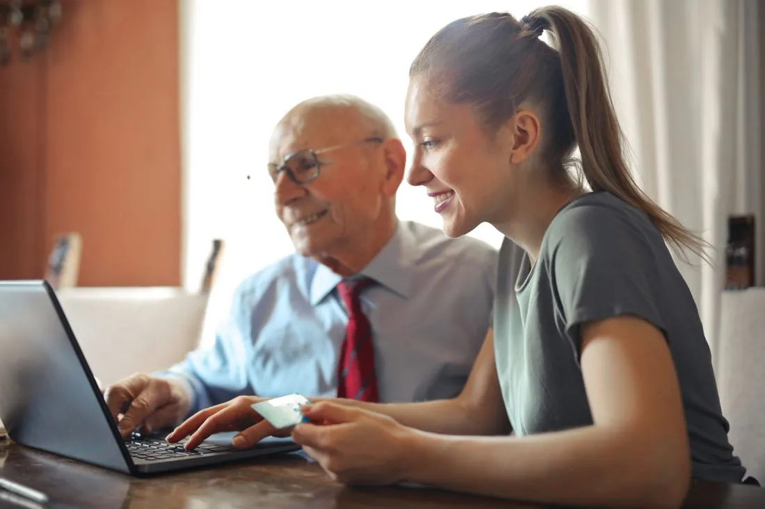 A carer sitting with an older man at home, helping him with a laptop