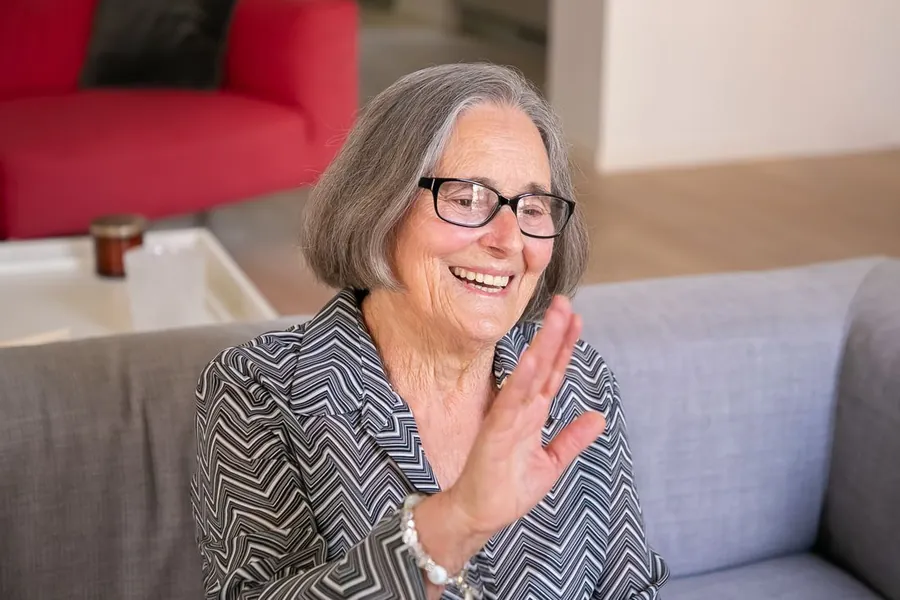 An older woman smiling and waving from her sofa at home